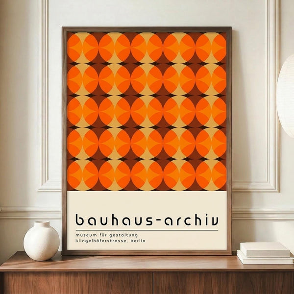 May include: Framed poster with geometric orange circular pattern in repeating grid layout against brown background, displayed on wooden credenza next to white ceramic vase and books in room with white paneled walls and neutral decor