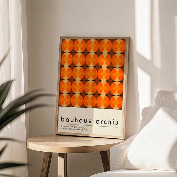 May include: Framed poster featuring a geometric pattern of orange and brown overlapping circles in a Bauhaus style, displayed on a light wood round side table next to a beige upholstered chair. Soft natural light streams across the minimalist white-walled room with a plant visible in the corner.