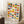 May include: Framed Bauhaus-style poster with colorful geometric circles and semicircles in yellow, orange, red, pink, blue, green, and teal arranged in an abstract pattern. The cream-colored print shows "BAUHAUS Est 1923" and "BAUHAUS AUSSTELLUNG WEIMAR JULI 1923" text at bottom. Wooden frame leans against white wall corner next to coffee cup on wooden stool and beige knit throw blanket on white textured rug.