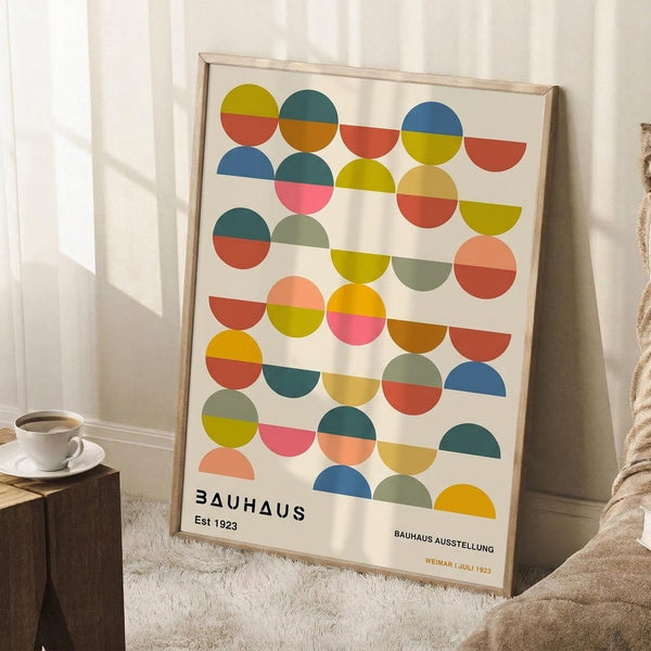 May include: Framed Bauhaus-style poster with colorful geometric circles and semicircles in yellow, orange, red, pink, blue, green, and teal arranged in an abstract pattern. The cream-colored print shows "BAUHAUS Est 1923" and "BAUHAUS AUSSTELLUNG WEIMAR JULI 1923" text at bottom. Wooden frame leans against white wall corner next to coffee cup on wooden stool and beige knit throw blanket on white textured rug.