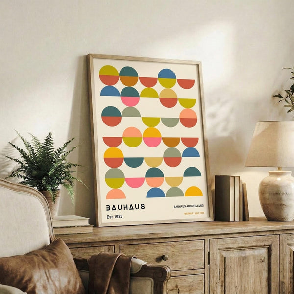 May include: Framed colorful geometric poster with overlapping semicircles in vibrant hues of yellow, orange, pink, blue, teal, and green on cream background, displayed on rustic wood sideboard alongside potted fern, ceramic vases, and books in cozy living room with beige armchair and neutral walls.