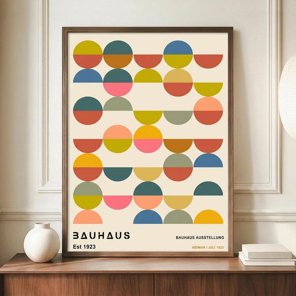 May include: Framed Bauhaus-style poster with geometric pattern of colorful half-circles and semicircles in yellow, blue, orange, pink, and green arranged in rows on cream background. Modern art print displayed on wooden sideboard next to white ceramic vase and stacked books in contemporary room with white paneled walls.