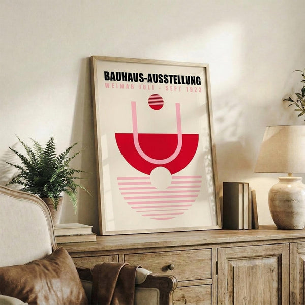 May include: Framed Bauhaus exhibition poster with geometric red and pink design displayed on rustic wooden sideboard in modern living room. Styled with fern plant, books, ceramic vase, and beige upholstered chair. Poster features bold typography and minimalist geometric shapes in red, pink and cream tones within light wood frame.