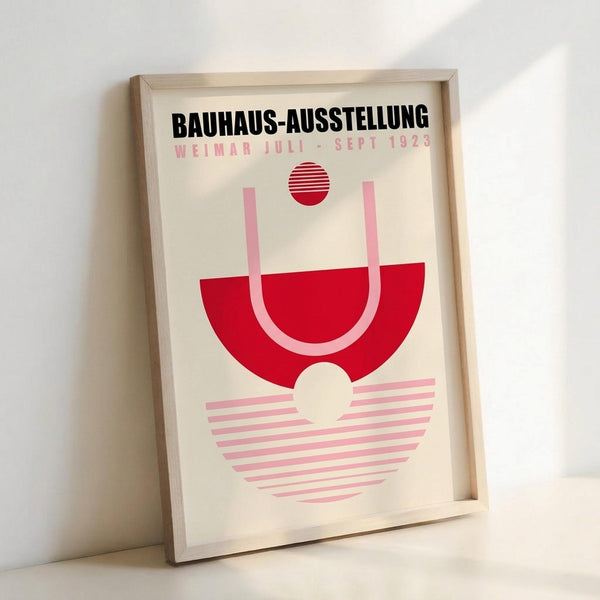 May include: Framed poster with geometric Bauhaus design featuring red semicircle, pink striped elements, and red striped circle on cream background. Light wood frame leaning against white wall, showing modern minimalist exhibition poster artwork in pink and red color palette.