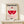 May include: Framed vintage-style Bauhaus exhibition poster in natural wood frame, leaning against white wall on light wooden shelf. Design features geometric shapes in red and pink - semicircle, circle, and striped elements on cream background with black and pink text. Small wrapped gift box with greenery sits in front of frame.