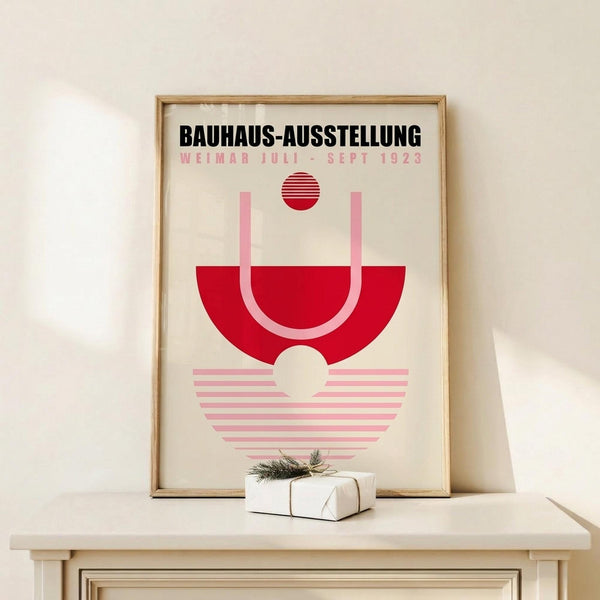 May include: Framed vintage-style Bauhaus exhibition poster in natural wood frame, leaning against white wall on light wooden shelf. Design features geometric shapes in red and pink - semicircle, circle, and striped elements on cream background with black and pink text. Small wrapped gift box with greenery sits in front of frame.