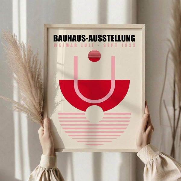 May include: Hands holding a framed geometric poster with red and pink shapes - a striped circle at bottom, red semi-circle in center, pink curved lines, and red striped circle at top. Black text reads "BAUHAUS-AUSSTELLUNG" and "WEIMAR JULI SEPT 1923" in pink. Light wood frame against white curtained window with dried pampas grass visible on left side.