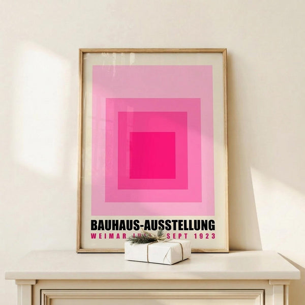May include: Framed poster with pink concentric squares creating an ombre effect from deep magenta center to pale pink outer edge, displayed on light wood shelf against cream wall. Small white wrapped gift with twine and evergreen sprig sits beside the thin wooden frame. The minimalist geometric design features bold black text below the squares.