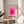 May include: Pink geometric poster with concentric squares in gradient shades from deep magenta center to pale pink edges, displayed in thin wooden frame on round wooden side table. White armchair and plant leaves visible in bright, minimalist room with natural lighting creating soft shadows on neutral walls.