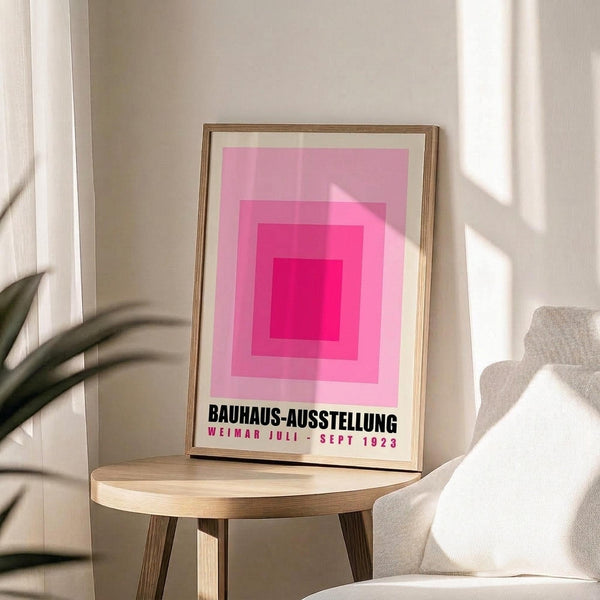 May include: Pink geometric poster with concentric squares in gradient shades from deep magenta center to pale pink edges, displayed in thin wooden frame on round wooden side table. White armchair and plant leaves visible in bright, minimalist room with natural lighting creating soft shadows on neutral walls.