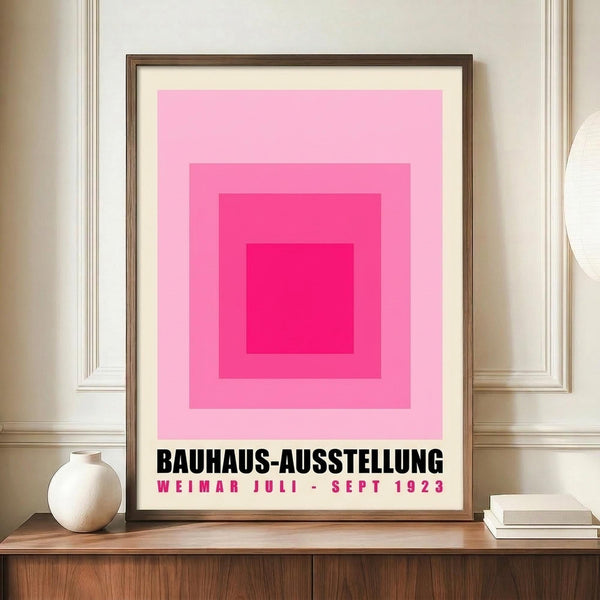 May include: Framed Bauhaus exhibition poster with concentric pink squares in graduating shades from deep magenta center to pale pink outer ring, displayed on wooden sideboard next to white ceramic vase and stacked books in elegant room with cream walls and white molding