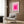 May include: Pink geometric Bauhaus exhibition poster with concentric squares in a light wood frame, displayed above a beige upholstered armchair with geometric patterned pillow. Modern reading nook setting with brass floor lamp, dark round side table with open book, wood paneled wall, and sheer curtains in natural lighting.