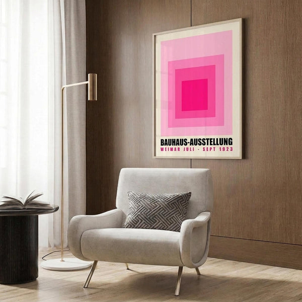 May include: Pink geometric Bauhaus exhibition poster with concentric squares in a light wood frame, displayed above a beige upholstered armchair with geometric patterned pillow. Modern reading nook setting with brass floor lamp, dark round side table with open book, wood paneled wall, and sheer curtains in natural lighting.