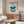 May include: Bauhaus exhibition poster from 1923 displayed above wooden sideboard in modern living room. Poster features geometric design with blue semicircle, black stripes, and cream background in thin frame. Room includes white curved chair, glass oval coffee table, and white table lamp. Clean minimalist interior with neutral tones and natural lighting.