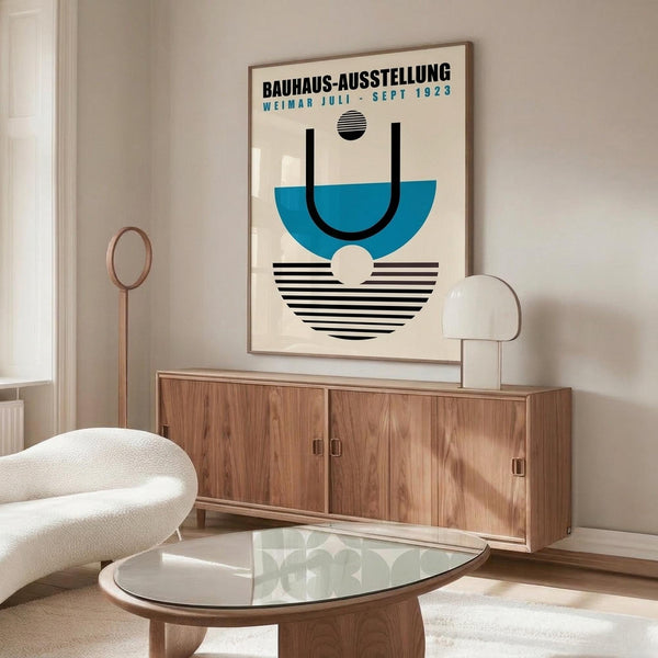 May include: Bauhaus exhibition poster from 1923 displayed above wooden sideboard in modern living room. Poster features geometric design with blue semicircle, black stripes, and cream background in thin frame. Room includes white curved chair, glass oval coffee table, and white table lamp. Clean minimalist interior with neutral tones and natural lighting.