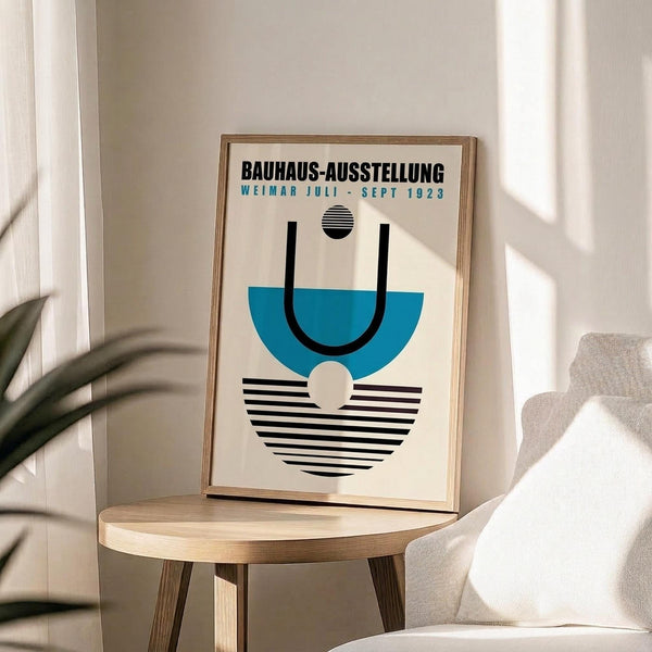 May include: Framed Bauhaus exhibition poster displayed on a light wood round side table next to a beige upholstered chair. The poster features geometric shapes in blue, black and white with striped patterns, housed in a natural wood frame. Soft natural lighting creates shadows across the minimalist white room setting with a plant partially visible in the corner.