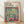 May include: Framed Henri Matisse exhibition poster featuring colorful painting of open French doors revealing tropical garden view, with teacup and potted plant on pink table in foreground. Vibrant blues, greens, oranges and reds throughout. Light wooden frame displayed against white wall on wooden floor with natural shadows.