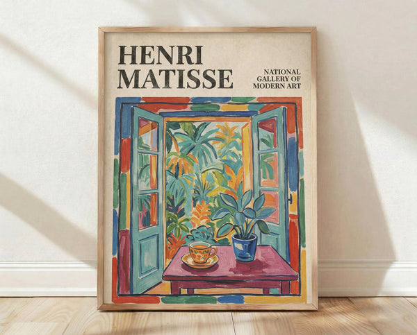 May include: Framed Henri Matisse exhibition poster featuring colorful painting of open French doors revealing tropical garden view, with teacup and potted plant on pink table in foreground. Vibrant blues, greens, oranges and reds throughout. Light wooden frame displayed against white wall on wooden floor with natural shadows.