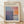 May include: Framed exhibition poster for Mark Rothko artwork displayed on light wooden floor against white wall with natural lighting creating shadows. The poster features abstract colorfield painting with blue, purple, orange and cream rectangular forms. Text shows gallery details and dates April 15 - July 30, 2023. Natural wood frame surrounds cream-colored poster on textured hardwood flooring.