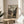 May include: Framed anime poster displayed on light wooden round table beside beige upholstered chair and plant. Poster shows two samurai characters sword fighting in traditional Japanese village setting with mountains in background. Earth-toned artwork in thin wooden frame catches natural sunlight streaming through white curtains in minimalist room.