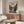 May include: Framed anime poster displayed above wooden mid-century modern credenza in minimalist living room. Poster shows two samurai characters dueling with swords in traditional Japanese village setting, featuring muted earth tones and Japanese text. Room has white upholstered chair, glass coffee table, modern floor lamp, and neutral beige walls with natural lighting.