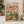 May include: Framed art print showing two white ducks in colorful Hawaiian shirts having a picnic, with one wearing sunglasses and the other a straw hat. The illustration features a pink background with confetti, green picnic table with bread basket, cheese board, wine bottles labeled "Quack Attack" and "Pond Prosecco," and small rubber ducks. The title "Duck Picnic" appears in multicolored letters at the top. Displayed in light wood frame on marble surface next to potted plant