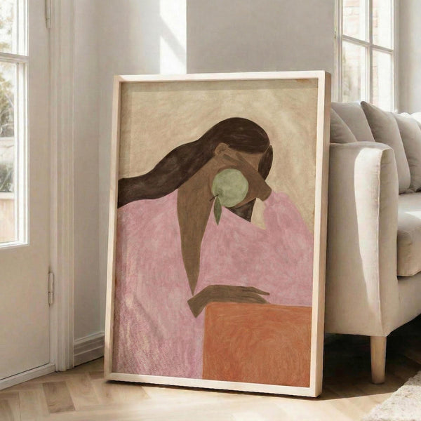 May include: Framed artwork leaning against white wall next to beige sofa in bright room with large windows. The poster shows a woman with long dark hair in pink clothing holding a green apple, painted in soft earth tones with textured brushwork. Light wood frame, approximately 2-3 feet tall, positioned on hardwood floor with natural sunlight streaming across the scene.