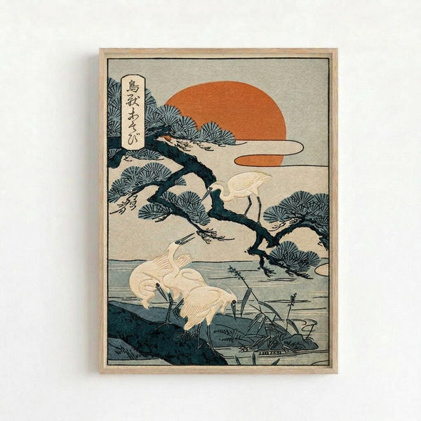 May include: Japanese woodblock print artwork displayed in light wood frame on white wall, featuring white cranes or herons among pine trees beside water, with orange sun in background and Japanese text in upper left corner, rendered in traditional ukiyo-e style with muted colors of teal, orange, and cream