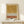 May include: Abstract watercolor artwork in wooden frame showing ochre yellow wash at top, dotted pattern in middle, and terracotta orange stripe below, displayed on light wood console table next to small wrapped gift with evergreen sprig against cream wall