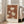 May include: Framed botanical art print leaning against white wall next to beige sofa in bright living room with large windows. The poster features stylized flowers and leaves in warm brown, cream and golden tones with text at bottom. Natural wood frame, approximately 2-3 feet tall, casting soft shadows on light wooden floor.