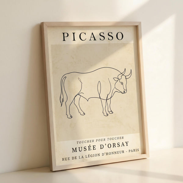 May include: Framed poster with cream background featuring minimalist black line drawing of a bull in Picasso's distinctive style. Text reads "PICASSO" at top and "TOUCHER POUR TOUCHER MUSÉE D'ORSAY RUE DE LA LÉGION D'HONNEUR - PARIS" at bottom. Light wooden frame displayed against white wall with soft natural lighting creating gentle shadows.