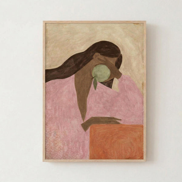 A framed artwork showing a woman with long dark hair in a pink garment, holding a green apple near her face. She appears to be looking down at an orange-red surface, possibly a table. The painting uses soft, muted colors with a beige background and has a minimalist, contemporary style. The piece is displayed in a light wood frame against a white wall.