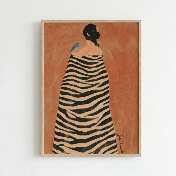 May include: Framed art print hanging on white wall showing woman from behind with dark hair in bun, wearing black and white striped cloak or blanket, small blue bird perched on her shoulder, warm orange-brown background, light wood frame, minimalist room setting