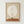May include: Framed surreal art print showing a small figure sitting on a curved brown hill beneath a large pale moon on beige background, displayed in natural wood frame against white wall. Minimalist composition with earthy tones and dreamlike scale contrast between tiny person and oversized celestial body.