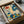 May include: Colorful floral art print in black frame, nestled in cardboard shipping box with tissue paper. The artwork features vibrant botanical illustration with red daisy, blue dotted flowers, yellow stems, and green foliage in folk art style on cream background. Box sits on white fabric surface next to rolled paper tube and dark glass object.
