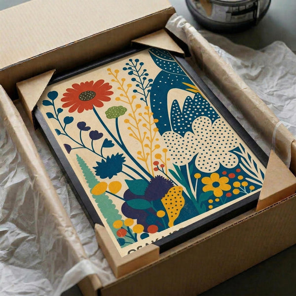 May include: Colorful floral art print in black frame, nestled in cardboard shipping box with tissue paper. The artwork features vibrant botanical illustration with red daisy, blue dotted flowers, yellow stems, and green foliage in folk art style on cream background. Box sits on white fabric surface next to rolled paper tube and dark glass object.
