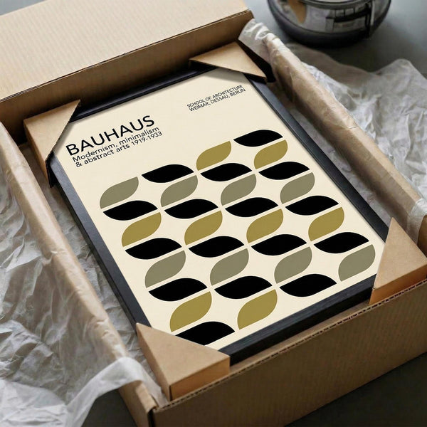 May include: Framed Bauhaus poster with geometric pattern of overlapping black and olive green oval shapes arranged in rows on cream background, being unpacked from cardboard shipping box with protective tissue paper wrapping on wooden surface