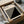 May include: A framed Bauhaus poster featuring bold black geometric shapes - diagonal stripes transitioning into a solid triangle - on cream background with minimalist typography. The black-framed print is nestled in brown cardboard packaging with white tissue paper, alongside wooden dowels, creating an unboxing scene on a neutral surface.