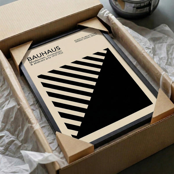 May include: A framed Bauhaus poster featuring bold black geometric shapes - diagonal stripes transitioning into a solid triangle - on cream background with minimalist typography. The black-framed print is nestled in brown cardboard packaging with white tissue paper, alongside wooden dowels, creating an unboxing scene on a neutral surface.