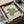 May include: Framed Bauhaus poster with green geometric design featuring flowing linear patterns forming abstract shapes on cream background, nestled in cardboard shipping box with protective tissue paper, wooden frame corners visible, sitting on white fabric surface next to cylindrical glass object