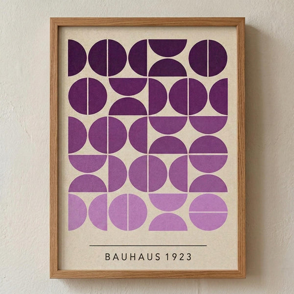 May include: Framed geometric art print with purple gradient circles and semicircles arranged in a grid pattern on cream background, displayed in light wood frame against textured white wall. Design features varying shades from deep purple to lavender with "BAUHAUS 1923" text below the geometric pattern.