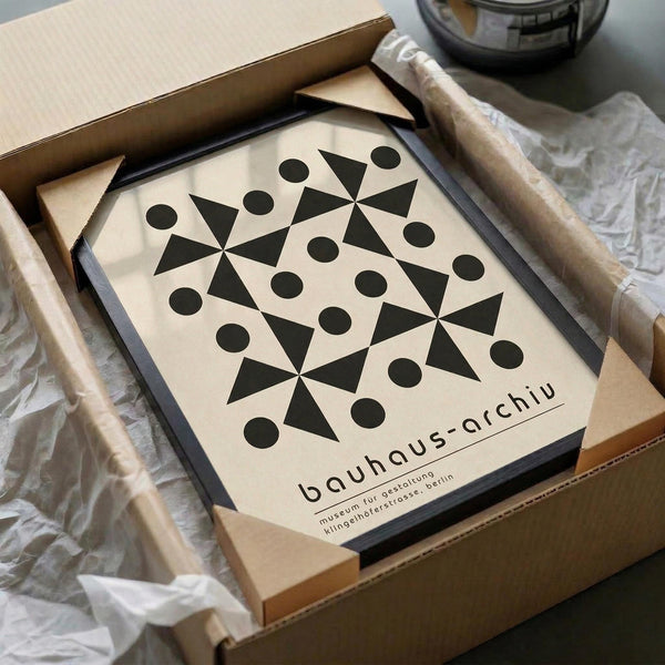 A geometric poster featuring black triangular shapes arranged in a symmetrical pattern with circles positioned around them on a cream background, displayed in a black frame. The poster is partially unpacked from brown cardboard packaging with white tissue paper, sitting on a light surface next to what appears to be a dark glass object.