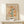 May include: Framed botanical poster with orange and yellow flowers on stems, surrounded by small leaves and botanical elements on a cream background, displayed in light wood frame on white wooden shelf next to small wrapped gift with twine and greenery sprig against neutral wall