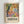 May include: Framed art poster displayed on white wall in light wooden frame. Colorful abstract painting features vibrant fruits, flowers, and foliage in Fauve style with bold yellows, oranges, greens, blues and pinks. Black tree branches frame organic shapes and botanical elements. White text below reads "DISCOVER THE ART OF HENRI MATISSE" with museum exhibition details in smaller print.