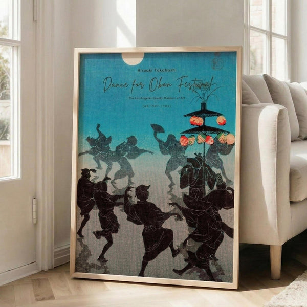 May include: Framed vintage Japanese woodblock print poster leaning against beige armchair in bright living room with white walls and large windows. Artwork features silhouetted figures in traditional dress performing folk dance under lanterns against turquoise sky background. Natural wood frame, approximately 2-3 feet tall, positioned on light wooden floor next to furniture.