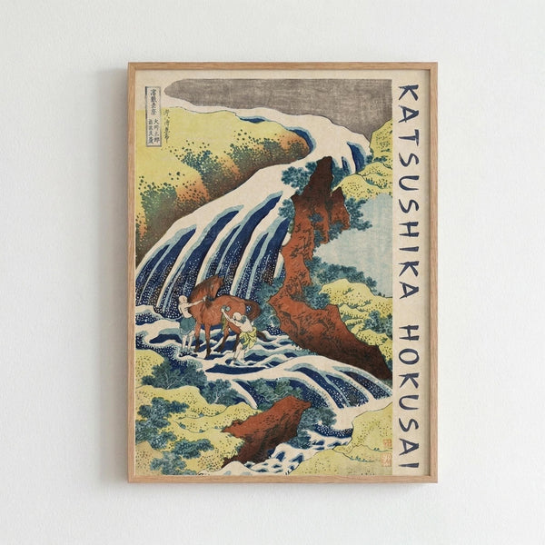 May include: Framed Japanese woodblock print showing a dramatic waterfall cascading over orange-brown rocky cliffs with white foam and blue water streams. Two small figures with a horse stand at the base of the falls. The artwork features traditional ukiyo-e style with muted yellows, greens, and blues depicting vegetation and landscape elements. Text reading "KATSUSHIKA HOKUSAI" appears vertically along the right edge. The print is displayed in a light wood frame against a white wall.