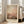 May include: Framed abstract art print leaning against beige sofa in bright living room with large windows. Artwork features soft orange, pink and brown tones with cloudy, atmospheric brushwork and artist name at bottom. Light wooden frame, approximately 2-3 feet tall, casting shadows on hardwood floor.