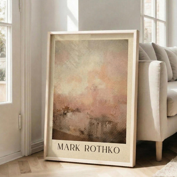 May include: Framed abstract art print leaning against beige sofa in bright living room with large windows. Artwork features soft orange, pink and brown tones with cloudy, atmospheric brushwork and artist name at bottom. Light wooden frame, approximately 2-3 feet tall, casting shadows on hardwood floor.