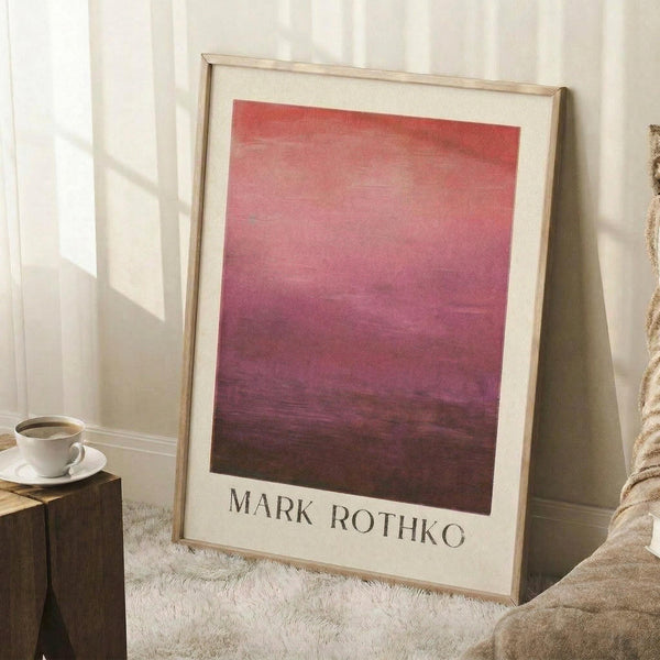 May include: Framed art print with gradient wash from coral red at top transitioning to deep purple and burgundy at bottom, displayed in light wooden frame with white matting. Print leans against white wall on fluffy white rug next to wooden side table holding white coffee cup and saucer, with beige textured cushion visible at edge.