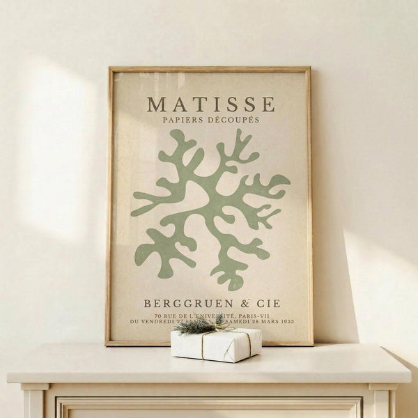 May include: Framed poster with cream background featuring sage green coral-like abstract cutout shape, displayed on light wood console table against white wall with small wrapped gift box beside it. Natural lighting creates soft shadows on the minimalist scene.